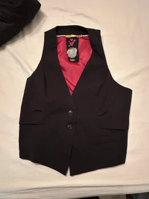 Svart väst med röd insida - Snygg svart väst från Iron Fist med två knappar framtill och cool röd insida. Västen har klassisk passform, två fickor fram och ett unikt innerfoder med mönster. Perfekt för att lyfta din outfit med en edgy touch.