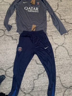 PSG träningsset Nike långärmad - Säljer ett PSG träningsset från Nike med långärmad grå tröja och mörkblå byxor. Tröjan har Qatar Airways-tryck och PSG-logga på bröstet. Byxorna har PSG-logga och Nike Swoosh. Materialet är lätt och andas, perfekt för träning.
