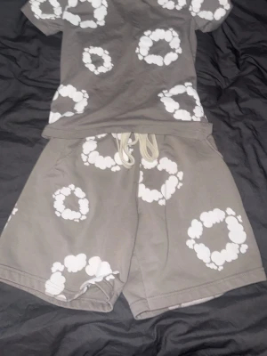 Grått set med vita molnringar - Säljer ett grått tvådelat set med t-shirt och shorts. Båda delarna har tryck av vita molnringar över hela plaggen. T-shirten har rund hals och korta ärmar, shortsen har resårmidja med snörning. Perfekt för en avslappnad och trendig stil.