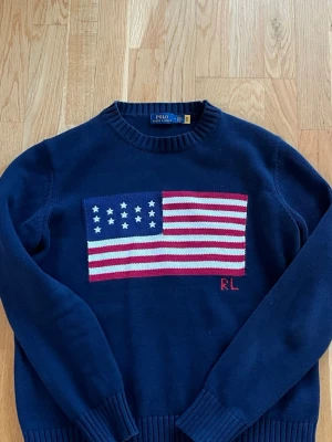 Mörkblå stickad tröja Polo Ralph Lauren - Mörkblå stickad tröja från Polo Ralph Lauren med amerikansk flagga i rött, vitt och blått på bröstet samt RL-broderi. Tröjan har rund halsringning och ribbstickade muddar vid ärmslut och nederkant. Perfekt för dig som gillar klassisk preppy stil.