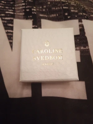 Smyckesask Caroline Svedbom - Vit smyckesask från Caroline Svedbom med guldfärgad logotyp och text på locket. Asken har en elegant och stilren design, perfekt för att förvara eller ge bort smycken. Materialet är präglat papper med lyxig känsla.