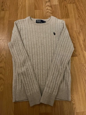Ljusgrå kabelstickad tröja Polo Ralph Lauren - Säljer en ljusgrå kabelstickad tröja från Polo Ralph Lauren med klassisk rund halsringning och svart broderad logga på bröstet. Tröjan har långa ärmar och ribbade muddar vid ärmslut och nederkant. Perfekt för dig som gillar stilren och tidlös design.