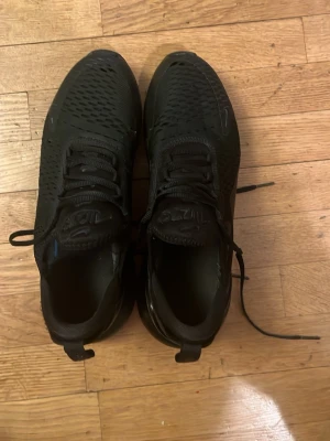 Nike Air Max 270 helsvarta sneakers - Snygga helsvarta Nike Air Max 270 sneakers med mesh-överdel och synlig Air-enhet i hälen. Skorna har klassisk snörning, diskret Swoosh-logga och en sportig, modern siluett. Perfekta för dig som gillar stilrena och bekväma sneakers. Lite synliga hål på framsidan men annars funkar de perfekt.
