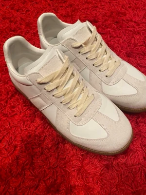 Vita sneakers med beige detaljer maison margiela - Säljer ett par stilrena vita sneakers med beige mockadetaljer och matchande beigea skosnören. Skorna har en klassisk siluett och gummisula i brunt. Perfekta för dig som gillar minimalistisk och clean stil. Maison margiela