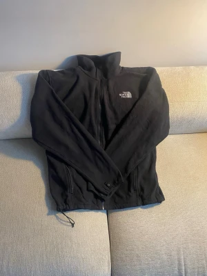 Svart fleecejacka från The North Face - Säljer en svart fleecejacka från The North Face med dragkedja framtill och hög krage. Jackan har två fickor med dragkedja och justerbar dragsko nedtill. Perfekt för lager-på-lager och outdoor-äventyr.