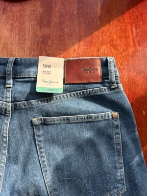 Blå tapered jeans från Pepe Jeans - Säljer ett par klassiska blå tapered jeans från Pepe Jeans London. Jeansen har normalhög midja, raka ben och är tillverkade i slitstarkt denim. Snygg läderpatch med logga baktill i midjan. Perfekta för en avslappnad och trendig look. 32W 34L