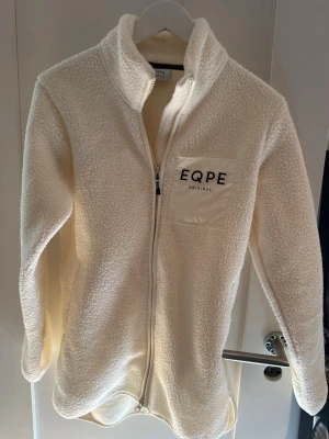 Vit teddyjacka från EQPE, M - Mysig vit teddyjacka från EQPE med dragkedja framtill och hög krage. Jackan har en bröstficka med EQPE-logga och är gjord i fluffigt teddy-material som håller dig varm. Perfekt för lager på lager och chill stil.