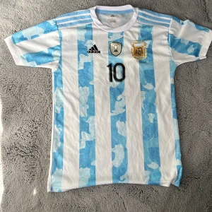 Argentina fotbollströja Messi 10 - Säljer en officiell Argentina fotbollströja från Adidas med Messi och nummer 10 på ryggen. Tröjan är vit med ljusblå ränder, har korta ärmar och broderade klubbmärken på bröstet. Perfekt för dig som älskar fotboll och Messi!
