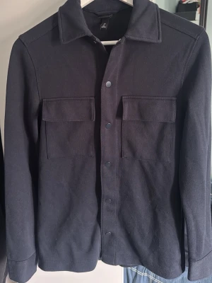 Svart overshirt med fickor - Stilren svart overshirt med två stora bröstfickor och tryckknappar framtill. Skjortkrage och långa ärmar med knapp vid ärmslut. Tillverkad i ett kraftigare tyg som ger en snygg och avslappnad look. Perfekt att bära öppen eller stängd över en t-shirt.