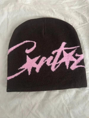 Brun mössa från Crittz med rosa text - Säljer en brun stickad mössa från Crittz med stor rosa text och stjärndetaljer framtill. Mössan har en klassisk form och är tillverkad i mjukt material som sitter skönt på huvudet. Perfekt för dig som vill ha en unik accessoar med streetwear-vibe.