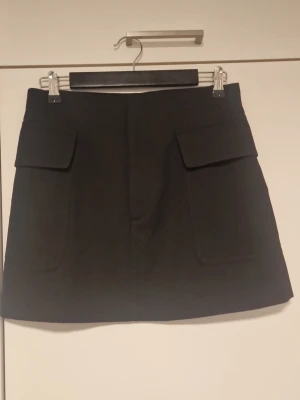 Svart kortkjol med fickor - Säljer en svart kortkjol med två stora fickor framtill. Kjolen har en enkel och stilren design som passar till det mesta. Perfekt att matcha med en stickad tröja eller en cool t-shirt.