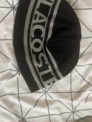 Svart mössa från Lacoste - Säljer en svart mössa från Lacoste med grått, brett logotypband runt hela mössan. Klassisk beanie-modell i mjukt stickat material som sitter skönt på huvudet. Perfekt för dig som gillar streetwear och vill ha en stilren accessoar.