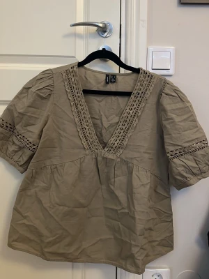Khaki grön blus från Vero Moda  - Snygg khaki grön blus från Vero Moda med djup V-ringning och virkat mönster längs halsringningen och på ärmarna. Blusen har puffiga korta ärmar och är tillverkad i ett lätt bomullstyg/linnetyg. Använd 1 gång! 💕nypris 499kr