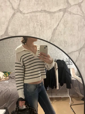 Vit stickad tröja med marinblå ränder - Trendig stickad tröja i vitt med marinblå horisontella ränder. Tröjan har en avslappnad passform, bred krage och långa ärmar. Perfekt till jeans för en casual look. Materialet ser ut att vara mjukt och bekvämt, troligen bomull eller bomullsblandning.