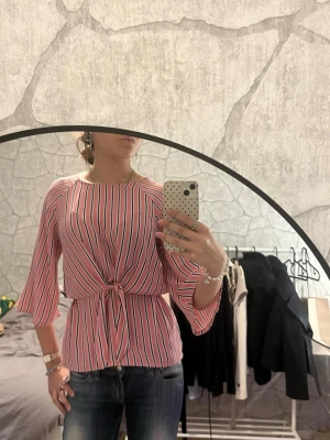 Rosa randig blus med knytning - Trendig rosa blus med vita och svarta vertikala ränder. Blusen har trekvartsärmar och en snygg knytning i midjan som ger en feminin siluett. Tillverkad i ett lätt och luftigt material, perfekt för en avslappnad men stilsäker look.