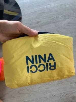 Gul handväska från Nina Ricci - Snygg gul handväska från Nina Ricci med blå logga på framsidan. Väskan har en mjuk, lätt och lite prasslig textil i ett enkelt format med dragkedja upptill. Perfekt för dig som gillar färg och vill ha något unikt i din väsksamling.