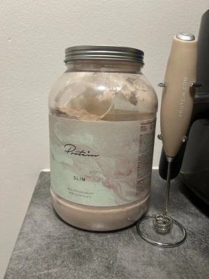 Protein Slim Mint Chocolate + Visp - Stor burk Protein Slim måltidsersättning med smak av mintchoklad från Protein.com. Medföljer en elektrisk visp i beige färg, perfekt för att blanda pulvret. Burken är genomskinlig med ett marmorerat etikettmönster i pastellfärger och har skruvlock i silver.