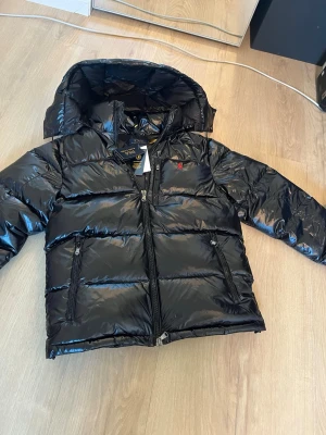 Svart dunjacka från Polo Ralph Lauren - Snygg svart dunjacka från Polo Ralph Lauren med blank finish, huva och två dragkedjeförsedda fickor. Jackan har RL-detalj på dragkedjan och den klassiska röda loggan broderad på bröstet. Perfekt för kalla vinterdagar.