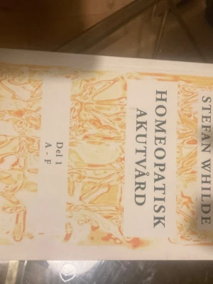 Homeopatisk Akutvård Del 1 A-F - Boken 'Homeopatisk Akutvård Del 1 A-F' av Stefan Whilde är en handbok inom homeopati. Omslaget är vitt med ett abstrakt mönster i gult och orange. Boken är tryckt på papper och har tydlig text på framsidan. Perfekt för dig som vill lära dig mer om alternativ medicin.