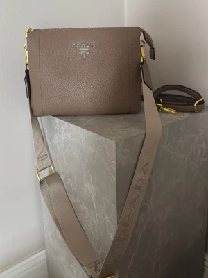 Beige axelväska från Prada - Snygg beige axelväska från Prada i grained läder med justerbar axelrem och guldfärgade metalldetaljer. Väskan har diskret logotyp framtill och en bred axelrem med präglad Prada-text. Perfekt för dig som gillar stilrena accessoarer. 23 cm bred