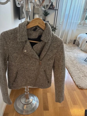 Grå ulljacka med dragkedja - Snygg grå jacka i ullmix med asymmetrisk dragkedja framtill och två snedställda fickor med dragkedja. Jackan har bred krage och lång ärm, perfekt för kyligare dagar. Enkel men trendig design som passar till det mesta.