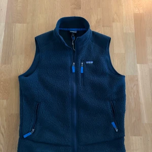 Patagonia väst - Mysig mörkblå fleeceväst från Patagonia med hög krage och tre fickor med dragkedja. Västen har en klassisk passform och är perfekt för lager-på-lager. Snygga blå detaljer på dragkedjorna och diskret logga på bröstet.