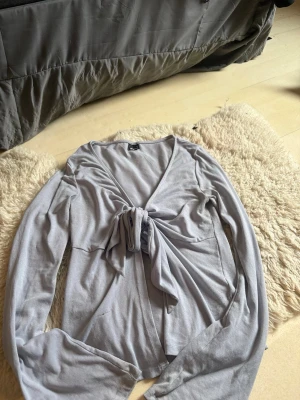 Ljusgrå omlottopp med knytband - Snygg ljusgrå omlottopp med långa ärmar och knytband framtill. Tillverkad i ett mjukt och stretchigt material som sitter skönt på kroppen. Perfekt att styla med jeans eller kjol för en chill look.