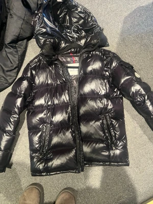 Moncler maya - Snygg svart dunjacka från Moncler med glansig finish och huva. Jackan har två dragkedjeförsedda fickor framtill och en Moncler-logga på ärmen. Perfekt för kalla vinterdagar tack vare det tjocka dunfodret och den klassiska puffade designen. Knappt använd, så i princip nyskick. Hör av er vid fler frågor🙌🏼