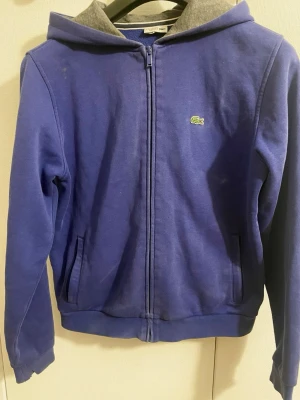 Blå zip hoodie från Lacoste - Snygg blå hoodie från Lacoste med dragkedja framtill och klassisk krokodil-logga på bröstet. Hoodien har huva med grått innerfoder och två fickor fram. Tillverkad i mjukt bomullsmaterial som är skönt att bära. Perfekt för en avslappnad och sportig stil. Denna kofta är lite större för att vara storlek 170 och motsvarar därmed storlek M.