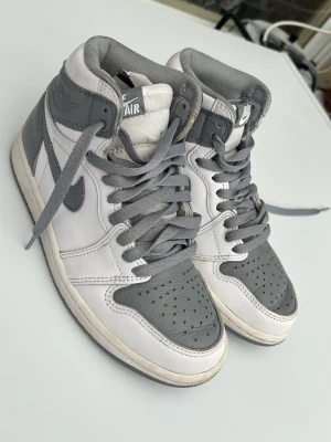 Nike Air Jordan 1 High grå/vit - Säljer ett par Nike Air Jordan 1 High sneakers i grått och vitt läder. Klassisk siluett med högt skaft, grå swoosh och matchande grå skosnören. Perforerad tåbox och ikoniska Air Jordan-loggan på sidan. Perfekta för dig som gillar streetwear och tidlös design.