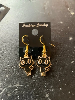 Svarta kattörhängen med gulddetaljer - Ett par söta örhängen med svarta kattmotiv och guldfärgade krokar. Katterna har stora ögon och en tecknad stil som ger en lekfull vibe. Materialet är metall med emalj i svart och guld. Perfekta för dig som gillar unika accessoarer med djurtema.