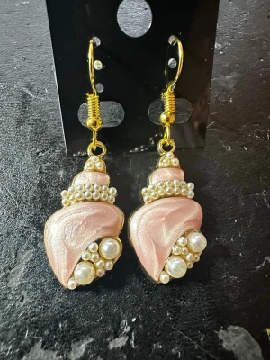 Rosa snäckformade örhängen med pärlor - Unika örhängen i snäckform med skimrande rosa yta och dekorerade med små vita pärlor. Krokarna är guldfärgade och ger en elegant touch. Perfekta för dig som vill sticka ut med ett kreativt och trendigt smycke.