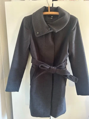 Mörkgrå kappa från H&M med knytskärp - Stilren mörkgrå kappa från H&M med bred krage och två knappar vid halsen. Kappan har ett matchande knytskärp i midjan och två sidofickor. Tillverkad i ett mjukt, ull-liknande material som håller dig varm under kyliga dagar. 💗