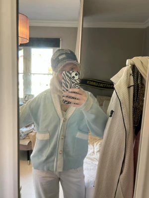 Ljusblå kofta med pärlknappar - Snygg ljusblå kofta med vita detaljer och pärlknappar. Koftan har två fickor framtill och ett mönstrat vitt kantband längs knäppningen och fickorna. Mjuk och skön, perfekt för lager-på-lager. Passar dig som gillar en söt och stilren look.