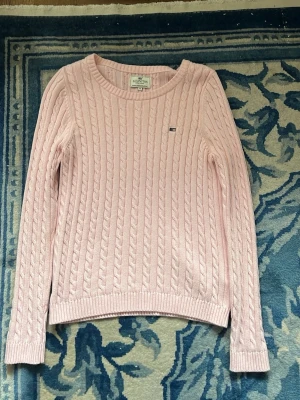 Ljusrosa kabelstickad tröja Lexington - Säljer en ljusrosa kabelstickad tröja från Lexington med rund halsringning och diskret logga på bröstet. Tröjan har långa ärmar och ribbade muddar vid ärmslut och nederkant. Perfekt för dig som gillar klassisk och stilren design.
