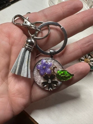 Nyckelring / väsksmycke med blomma och tofs - Snygg nyckelring / väsksmycke i metall med rund bricka dekorerad med en lila blomma, svart blomma och grön emaljerad blad. Tofs i ljusgrå mockaimitation ger extra stil. Perfekt att hänga på väskan eller nycklarna för en personlig touch.