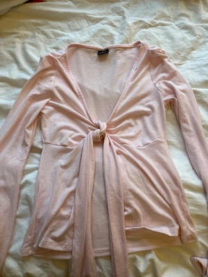 Ljusrosa omlottopp från Gina Tricot - Supersöt ljusrosa omlottopp från Gina Tricot med långa ärmar och knyt framtill. Toppen har en djup v-ringning och är gjord i mjuk bomull, perfekt för en trendig och avslappnad look.