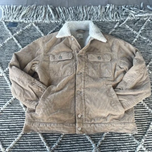 Beige manchesterjacka med teddykrage - Säljer en beige manchesterjacka med vit teddykrage och två bröstfickor med knappar. Jackan har knappstängning framtill och är fodrad för extra värme. Bra skick, använd och det ser man lite på insidan men det är inte som påverkan jackan.