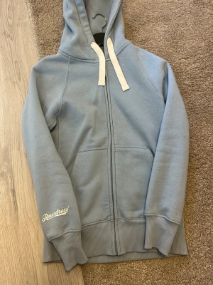Ljusblå zip hoodie från Russedress - Ljusblå hoodie med dragkedja från Rustdress. Tröjan har vit dragsko i huvan, ribbade muddar och fickor framtill. Perfekt för en avslappnad stil och enkel att matcha med jeans eller mjukisbyxor.