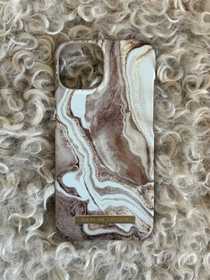 Marmorerat mobilskal från iDeal of Sweden IPhone 13 - Snyggt mobilskal med marmormönster i bruna, beige och vita toner från iDeal of Sweden. Skalet har en glansig finish och en guldfärgad logga längst ner. Passar perfekt för dig som vill ha ett trendigt och stilrent skydd till mobilen. 