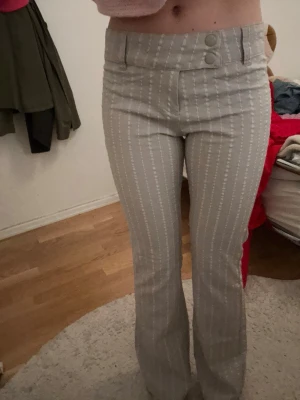 Grå kostymbyxor med vita ränder - Snygga grå kostymbyxor med vita vertikala ränder och två dekorativa knappar i midjan. Byxorna har hög midja och utsvängda ben för en trendig siluett. Perfekta för dig som vill ha en stilren och modern look.