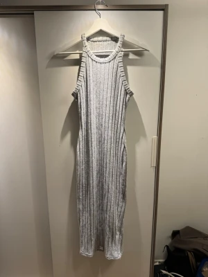 Grå ribbad långklänning - Snygg grå ribbad långklänning med ärmlös design och hög hals. Klänningen har en kroppsnära passform och är tillverkad i ett mjukt stickat material som ger en bekväm känsla. Perfekt för dig som gillar stilrena och moderna plagg.