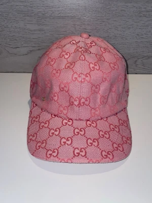 Rosa GG-keps från Gucci - Säljer en rosa keps från Gucci med det klassiska GG-mönstret över hela kepsen. Kepsen har böjd skärm och är tillverkad i ett slitstarkt tygmaterial. Perfekt för dig som vill sticka ut med en lyxig och trendig accessoar.