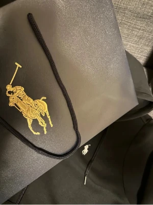 Svart hoodie från Polo Ralph Lauren - Snygg svart hoodie från Polo Ralph Lauren med dragkedja framtill och klassisk vit logga broderad på bröstet. Hoodien har justerbar huva med snören och praktiska fickor framtill. Perfekt för en avslappnad och stilren look.