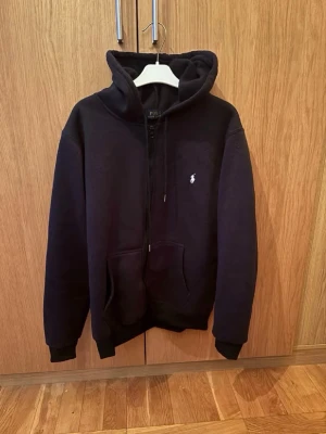 Svart hoodie från Polo Ralph Lauren - Klassisk svart hoodie från Polo Ralph Lauren med dragkedja framtill och vit broderad logga på bröstet. Hoodien har kängurufickor, ribbade muddar och en rejäl huva. Tillverkad i mjukt bomullsmaterial som är skönt att ha på sig.  Säljer åt en kompis som säljer pågrund av att han är för stor för den/ osäker på äkthet därav av ett skit bra pris tveka inte på att skriva om frågor eller om du har funderingar kan gå ner lite i pris vid snabb affär 