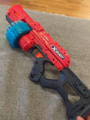 X-Shot leksakspistol med trumma - Cool leksakspistol från X-Shot i rött, svart och blått med stor roterande trumma för skumdart-pilar. Pistolen har futuristisk design och ett greppvänligt handtag. Perfekt för roliga dart-battles med vänner!