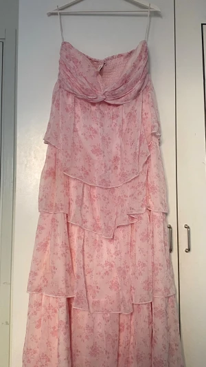 Rosa blommig långklänning från Nelly - Romantisk rosa långklänning från Nelly med blommigt mönster och volangdetaljer. Klänningen är axelbandslös och har flera lager i ett lätt, transparent material som ger en drömmig känsla. Perfekt för sommarens alla tillfällen. Använd endast 1 gång men medföljande sjalen är borttappad så säljer för rabatt  ORG pris: 900