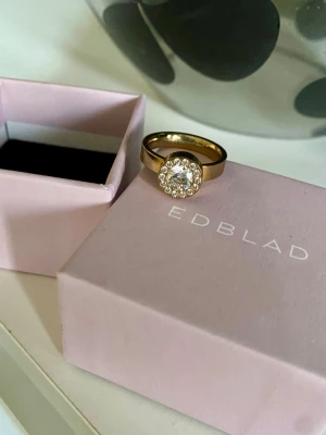 Edblad Thassos Ring Gold. Nyskick med presentask! - Snygg guldfärgad ring från Edblad, modell Thassos. Ringen har en rund, glittrande mittsten omgiven av små klara stenar som ger en lyxig känsla. Perfekt accessoar för att lyfta din stil. Materialet är rostfritt stål med guldfinish. Storlek M. Nyskick!