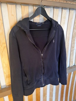 Navy cardigan/jacka från Polo Ralph Lauren - Säljer en navy cardigan/jacka från Polo Ralph Lauren med dragkedja och huva. Hoodien har fickor på sidorna och ribbade muddar vid ärmslut och nederkant. Perfekt för en avslappnad och stilren look.