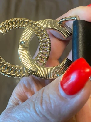 Svart Chanel bälte med gulddetaljer - Snyggt svart bälte från Chanel med ikoniskt dubbel-C-spänne i guld. Spännet har kedjedetaljer och texten CHANEL ingraverad. Perfekt accessoar för att lyfta vilken outfit som helst.